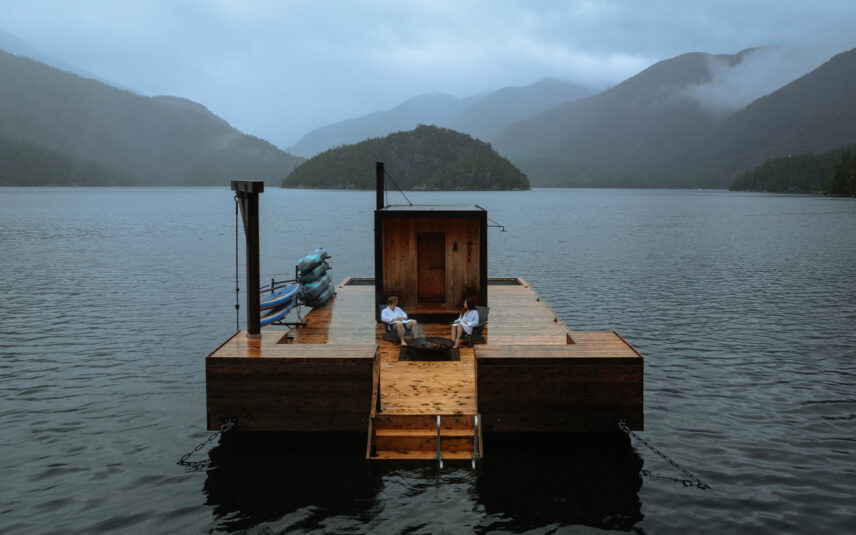 Couple in white robes relaxing by a fire pit at a floating sauna with mountains and morning fog in Tofino, BC