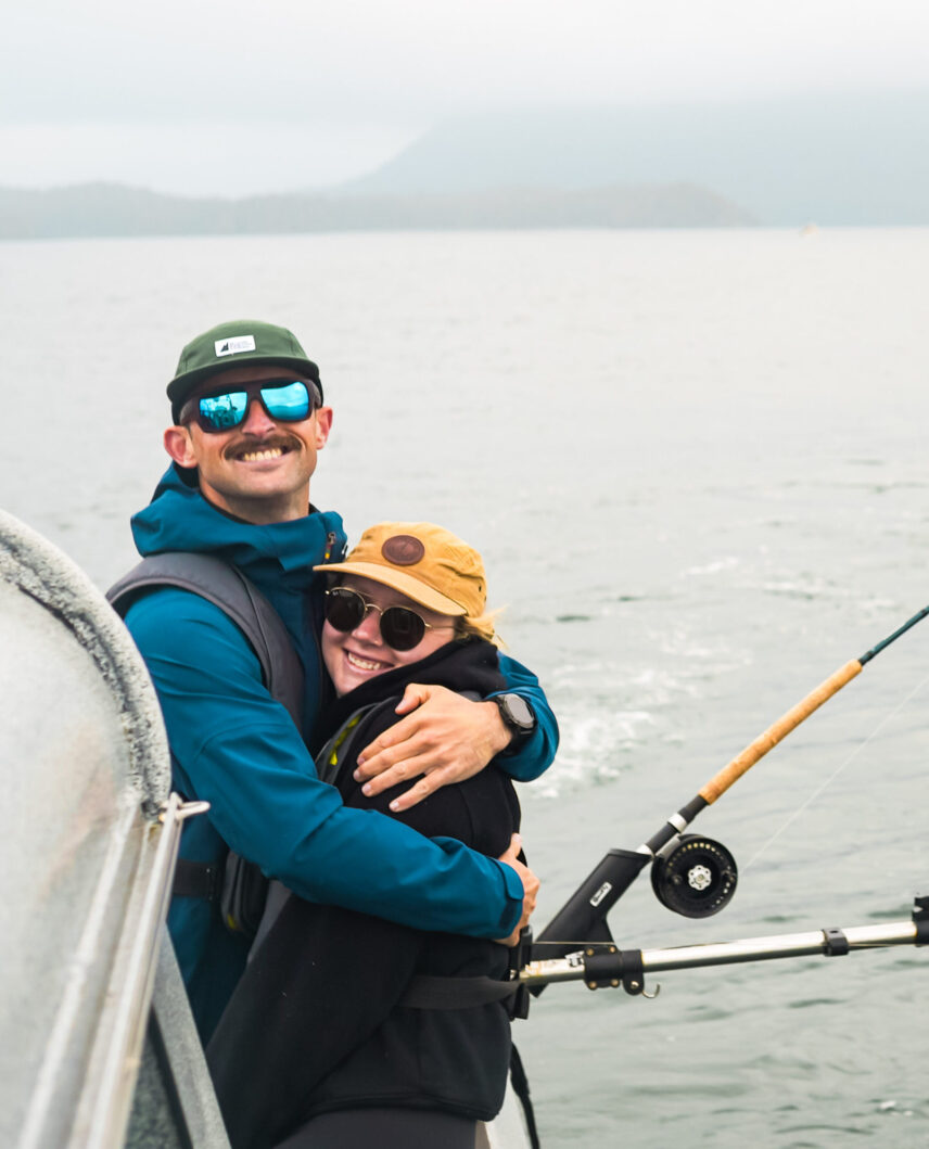 A couple hugging and smiling at the camera in a fishing boat with fishing line in the background