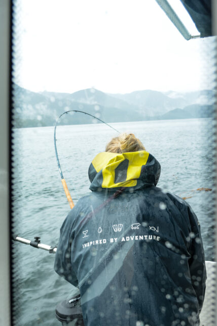 Reflection of a man fishing wearing a jacket that says 'inspired by adventure' with the TRM icons