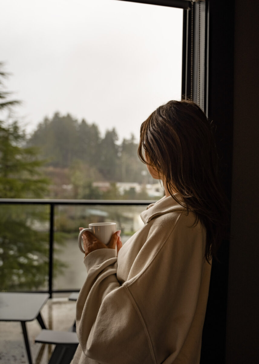 Woman looking out the window to the rainy outside while holding a warm drink